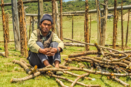 Beni, Nepal - circa May 2012: Native man with black knitted cap on his head sits on ground surrounded by wooden branches in Beni, Nepal. Documentary editorial.のeditorial素材