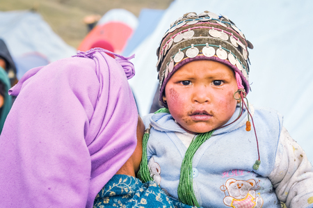 Dolpo, Nepal - circa May 2012: Small child in blue sweater with green necklace and cap with coins has dirt on face from arms of mother in Dolpo, Nepal. Documentary editorial.のeditorial素材