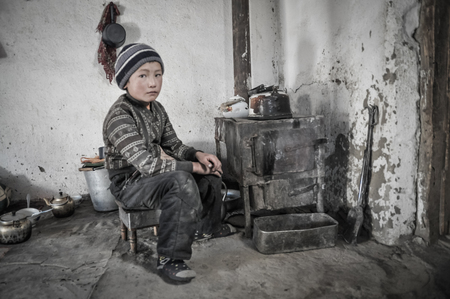 Karakul, Tajikistan - circa September 2011: Young boy sits on small chair next to small old furnace in Karakul, Tajikistan. Documentary editorial.のeditorial素材