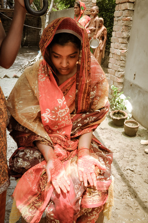 Paigacha, Bangladesh - circa July 2012: Native people with children stand in mud and girl in red pours water from jar on head of halfnaked sitting boy in Paigacha, Bangladesh. Documentary editorial.のeditorial素材