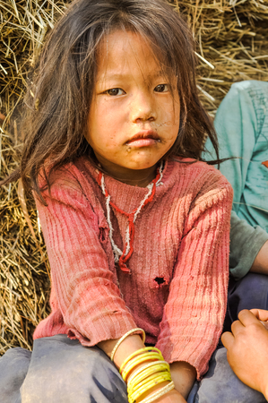 Kanchenjunga Trek, Nepal - circa April 2012: Young girl in pink sweater with hole in it sits on ground and looks sadly to photocamera in Kanchenjunga Trek, Nepal. Documentary editorial.のeditorial素材