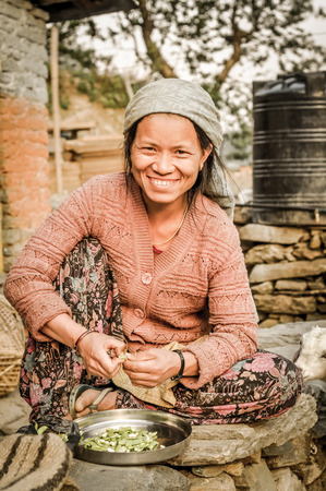 Beni, Nepal - circa May 2012: Smiling native woman with white headcloth and in pink sweater sits on ground and prepares meal in Beni, Nepal. Documentary editorial.のeditorial素材