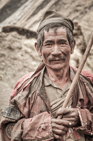 Dolpo, Nepal - circa May 2012: Old native man with grey moustache wears brown cap and brown clothes and holds wooden stick in his hands in Dolpo, Nepal. Documentary editorial.のeditorial素材