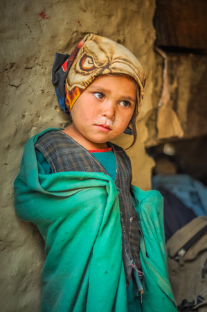 Beni, Nepal - circa May 2012: Young native girl with headcloth in green shirt and vest leans on wall and covers herself in green blanket in Beni, Nepal. Documentary editorial.のeditorial素材