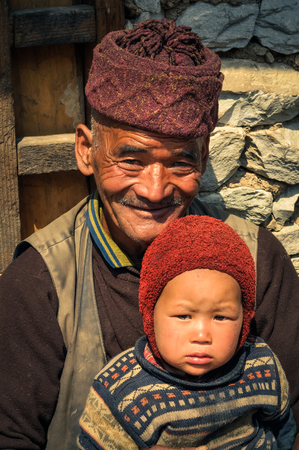 Dolpo, Nepal - circa May 2012: Smiling native man with red knitted cap with his grandchild on his knees in front of their house in Dolpo, Nepal. Documentary editorial.のeditorial素材