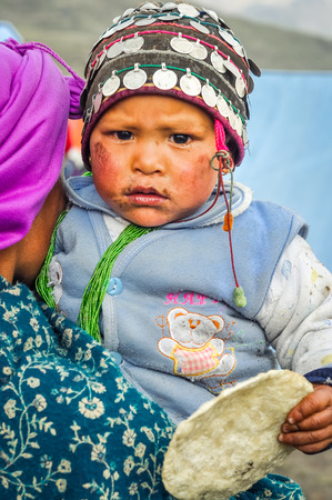 Dolpo, Nepal - circa May 2012: Small child in blue sweater with green necklace and cap with coins has dirt on face with cake in hand in Dolpo, Nepal. Documentary editorial.のeditorial素材