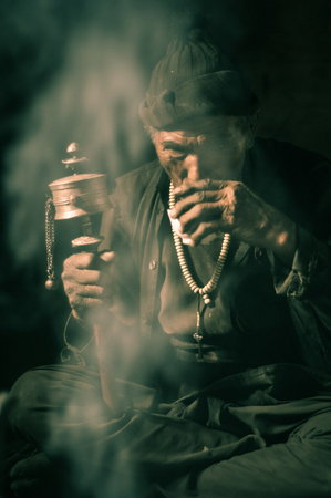 Dolpo, Nepal - circa May 2012: Old man dressed in brown shirt, vest and brown cap on head sits on ground and drinks covered in smoke in Dolpo, Nepal. Documentary editorial.のeditorial素材