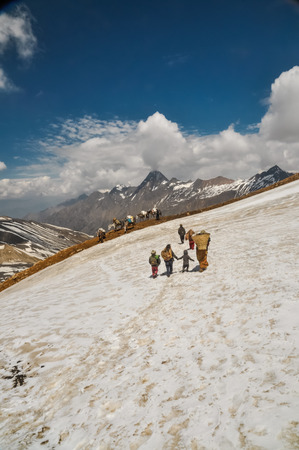 Dolpo, Nepal - circa June 2012: Native woman walks in snow with basket full of load and with her three small children in beautiful snowy mountains in Dolpo, Nepal. Documentary editorial.のeditorial素材