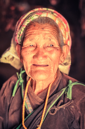 Dolpo, Nepal - circa May 2012: Old woman with grey hair and colourful headcloth dressed in brown shirt with yellow necklace made of beads smiles gently in Dolpo, Nepal. Documentary editorial.のeditorial素材