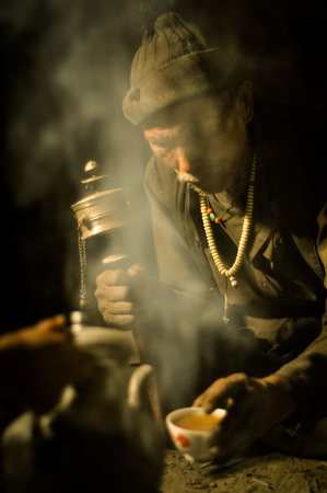 Dolpo, Nepal - circa May 2012: Old man with brown cap on head sits on ground and looks down to cup of tea covered in smoke in Dolpo, Nepal. Documentary editorial.のeditorial素材