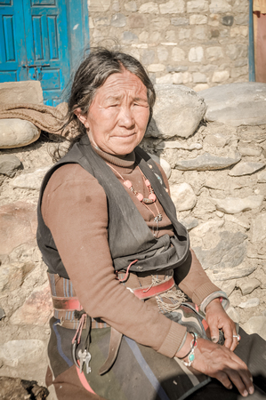 Dolpo, Nepal - circa June 2012: Old native woman with wrinkles on face dressed in brown shirt and black vest with orange necklace sits on stone in Dolpo, Nepal. Documentary editorial.のeditorial素材