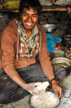 Dolpo, Nepal - circa May 2012: Smiling young man with black hair dressed in red shirt sits on ground and prepares meal in Dolpo, Nepal. Documentary editorial.のeditorial素材