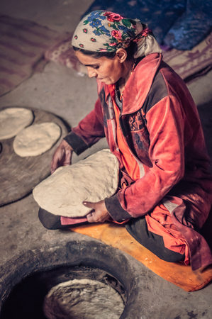 Wakhan valley, Tajikistan - circa October 2011: Woman in red clothes sits on ground and prepares meal from dough in Wakhan valley, Tajikistan. Documentary editorial.のeditorial素材
