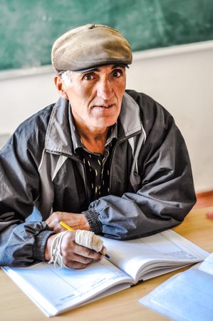 Khorog, Tajikistan - circa September 2011: Teacher with cap on his head sits at table and writes to exercise book at school in Khorog, Tajikistan. Documentary editorial.のeditorial素材
