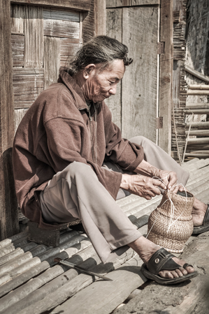 Ziro, Arunachal Pradesh - circa March 2012: Older man in brown shirt sits on ground and weaves basket in Ziro, Arunachal Pradesh. Documentary editorial.のeditorial素材