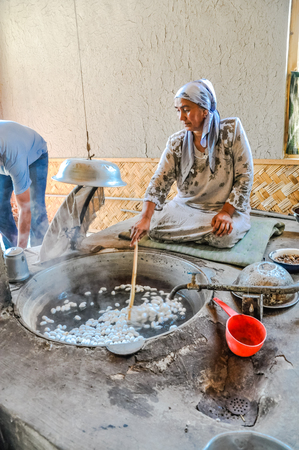 Margilan, Uzbekistan - circa July 2011: Woman sits on ground and stirs silk worm cocoons with wooden stick in silk factory in Margilan. Documentary editorial.のeditorial素材
