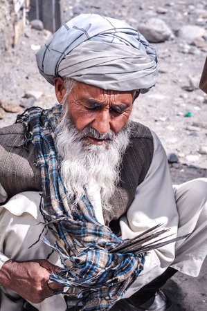 Ishkoshim, Tajikistan - circa October 2011: Older man with turban on his head wipes sticks with cloth at market in Ishkoshim, Tajikistan. Documentary editorial.のeditorial素材
