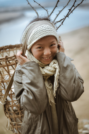 Menchuka, Arunachal Pradesh - circa March 2012: Smiling young girl wears grey jacket and carries basket on her back with help of her head in Menchuka, Arunachal Pradesh. Documentary editorial.のeditorial素材