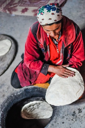 Wakhan valley, Tajikistan - circa October 2011: Woman in red clothes and with headcloth sits on ground and prepares flat cakes from dough in Wakhan valley, Tajikistan. Documentary editorial.のeditorial素材