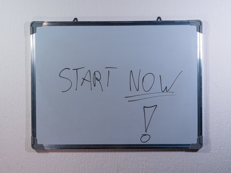 Whiteboard showing START NOW message on clean white wall background representing motivation, action and new beginningの写真素材