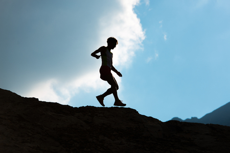 Woman runs downhill on mountain trail in silhouette against the skyの写真素材