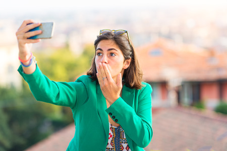 Young beautiful girl taking sends a kiss away selfies with mobile phone in the cityの写真素材