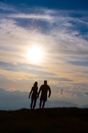 Couple in silhouette holding hands walking towards colorful clouds at sunset in the mountains.の写真素材