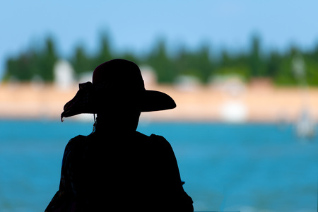 Silhouette of tourist with sun hat looks at the Venetian lagoon in blurred background.の写真素材
