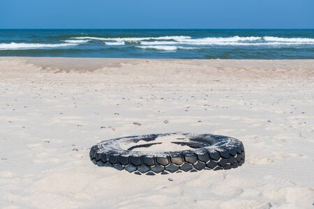 Big black rubber tire left on a sandy beach with blue sea and waves on background, environment pollution conceptの写真素材