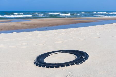 Big black rubber tire left on a sandy beach with blue sea and waves on background, environment pollution conceptの写真素材