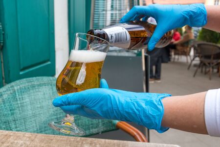 Hands of waiter or waitress dressing plastic gloves serving beer to the client at the table of an outdoor bar, cafe or restaurant, reopening after quarantine restrictionsの写真素材