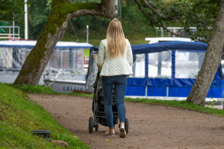 Blonde hair young mother with baby in a stroller walking in a park by a lake