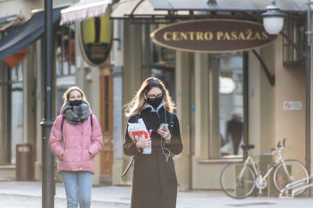 Vilnius, Lithuania - November 22 2020: Girl with phone wearing glasses and protective face mask walking in the city near a shop or shopping center during Covid or Coronavirus outbreakのeditorial素材