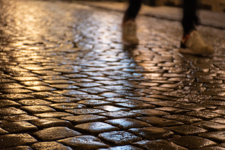 Wet cobblestones at night after rain on a street or road surface, bottom view with shadow or silhouette of legs and feet of a person walking or runningの写真素材