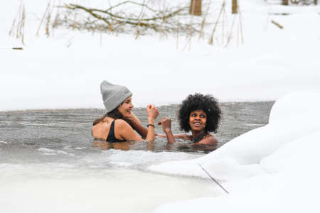 Couple of beautiful girls enjoying winter and swimming in the cold water of a lake or river, cold therapy, ice swim with white winter landscape and forest on backgroundのeditorial素材