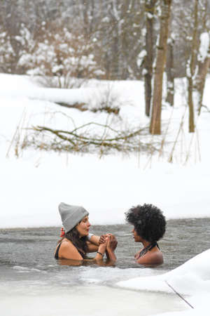 Couple of beautiful girls enjoying winter and swimming in the cold water of a lake or river, cold therapy, ice swim with white winter landscape and forest on background, verticalのeditorial素材