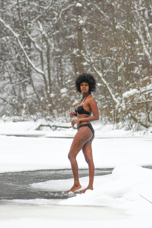 Beautiful girl on the ice and enjoying winter in the cold water of a lake or river, cold therapy, ice swim with white winter landscape and forest on background, verticalのeditorial素材