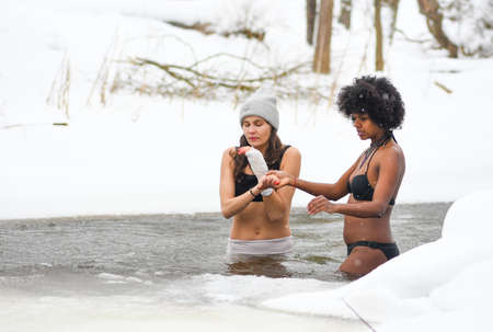 Couple of beautiful girls enjoying winter and swimming in the cold water of a lake or river, cold therapy, ice swim with white winter landscape and forest on backgroundのeditorial素材