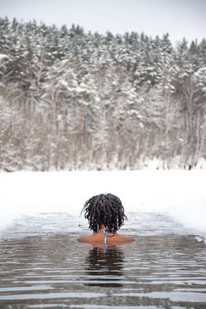 Beautiful girl swimming in the cold water of a lake or river, cold therapy, ice swim with winter landscape and forest on background, verticalの写真素材