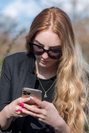 Vilnius, Lithuania - April 2021: Beautiful young blonde hair girl with sunglasses checking the smartphone in the street, verticalのeditorial素材