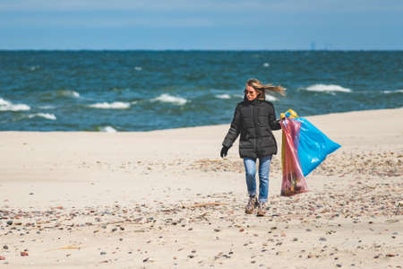Beautiful blond girl collecting plastic trash rubbish garbage on the sand beach and putting it into...