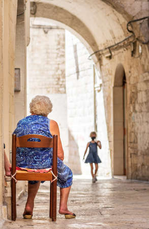 Grandmother waiting for her granddaughter on the doorstep in the narrow streets of Bari old town, Puglia, Italy, verticalの写真素材