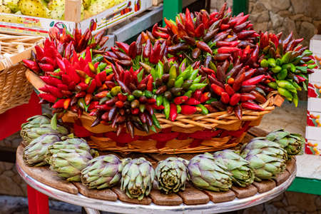 Fresh green artichokes with green leaves and bunches of red and green hot chili peppers in a basket in a street food marketの写真素材