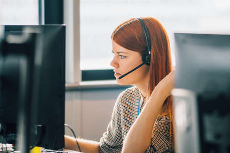 Beautiful young Caucasian red hair girl dressing casual working in the office or class at the desk in front of two monitors. Focus on job, concentration and study conceptの写真素材