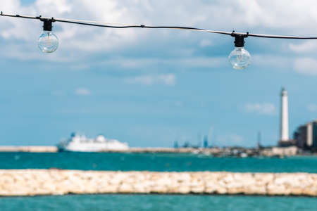 Vintage light bulbs with blue sky, white clouds, blue sea and breakwater, Bari port or harbor with ships and lighthouse on blurred backgroundの写真素材