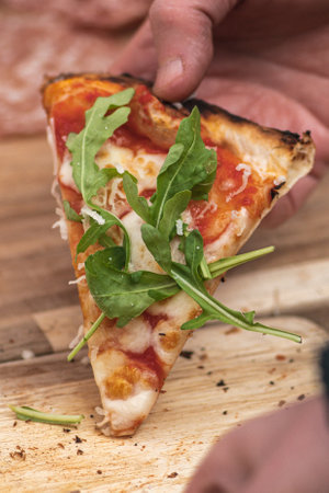 Hand taking last piece or slice of pizza with tomato sauce, mozzarella and fresh rocket leaves from...