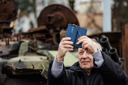 Kyiv, Ukraine - February 25 2023: Elderly man taking a selfie near a burnt and melted rusty wreckage of a Soviet Russian-made tank T-72B destroyed during Russian invasion of Ukraine in Kievのeditorial素材