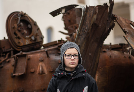 Kyiv, Ukraine - February 25 2023: Child photographed near a burnt and melted rusty wreckage of a Soviet Russian-made tank T-72B destroyed during the Russian invasion of Ukraine in a square in Kievのeditorial素材