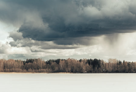Beautiful early spring landscape with frozen lake, forest on the horizon and dramatic sky with gray...