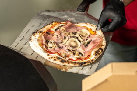 A person wearing black gloves is carefully lifting a freshly baked pizza from an electric oven using a pizza peelの写真素材